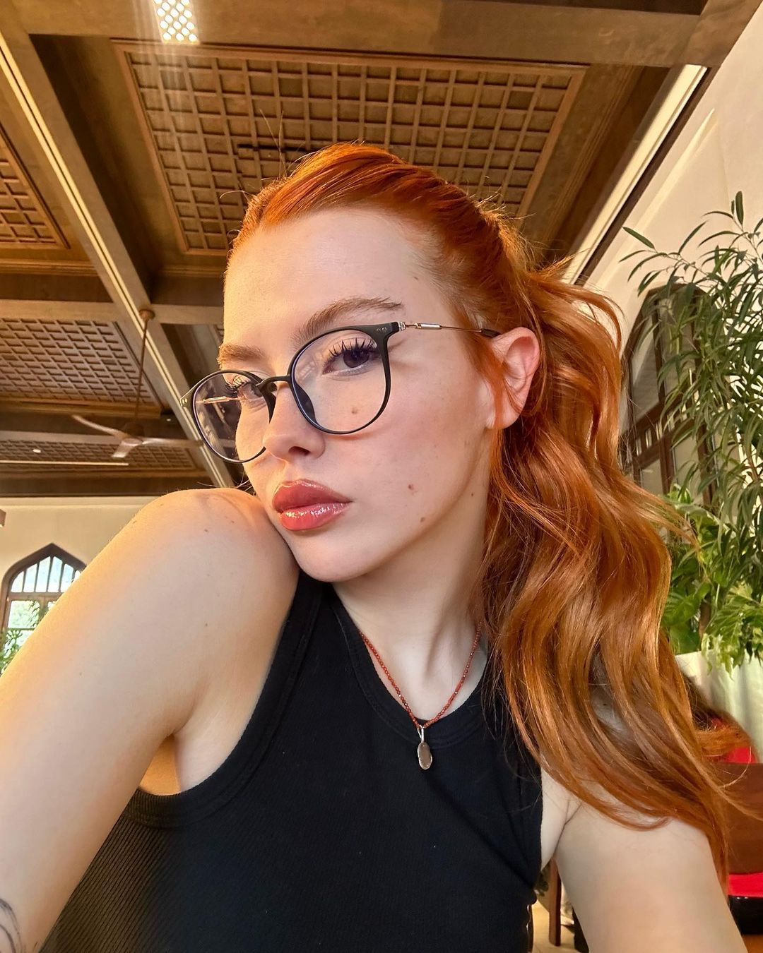 A woman sporting vibrant red hair, glasses, and a black top, exuding a confident and artistic vibe.