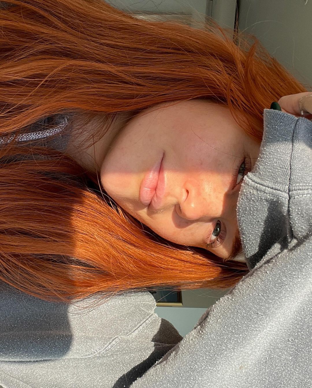 A person with striking red hair partially covering their face with a grey sleeve, accented by sunlight