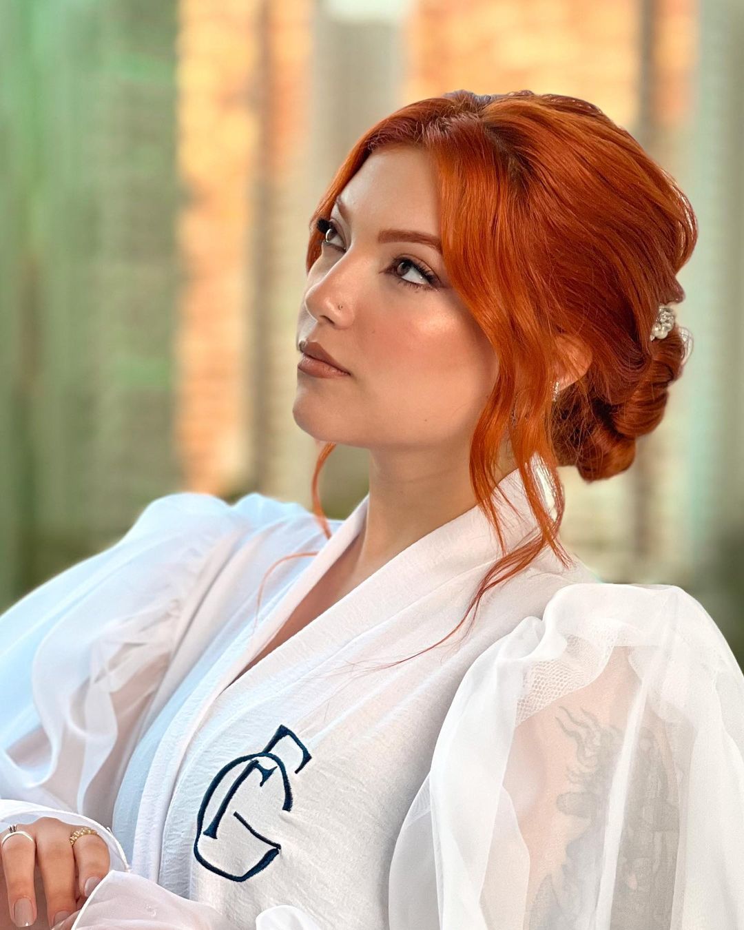 A person with red hair styled in an elegant updo, wearing a sheer white robe with monogrammed initials.
