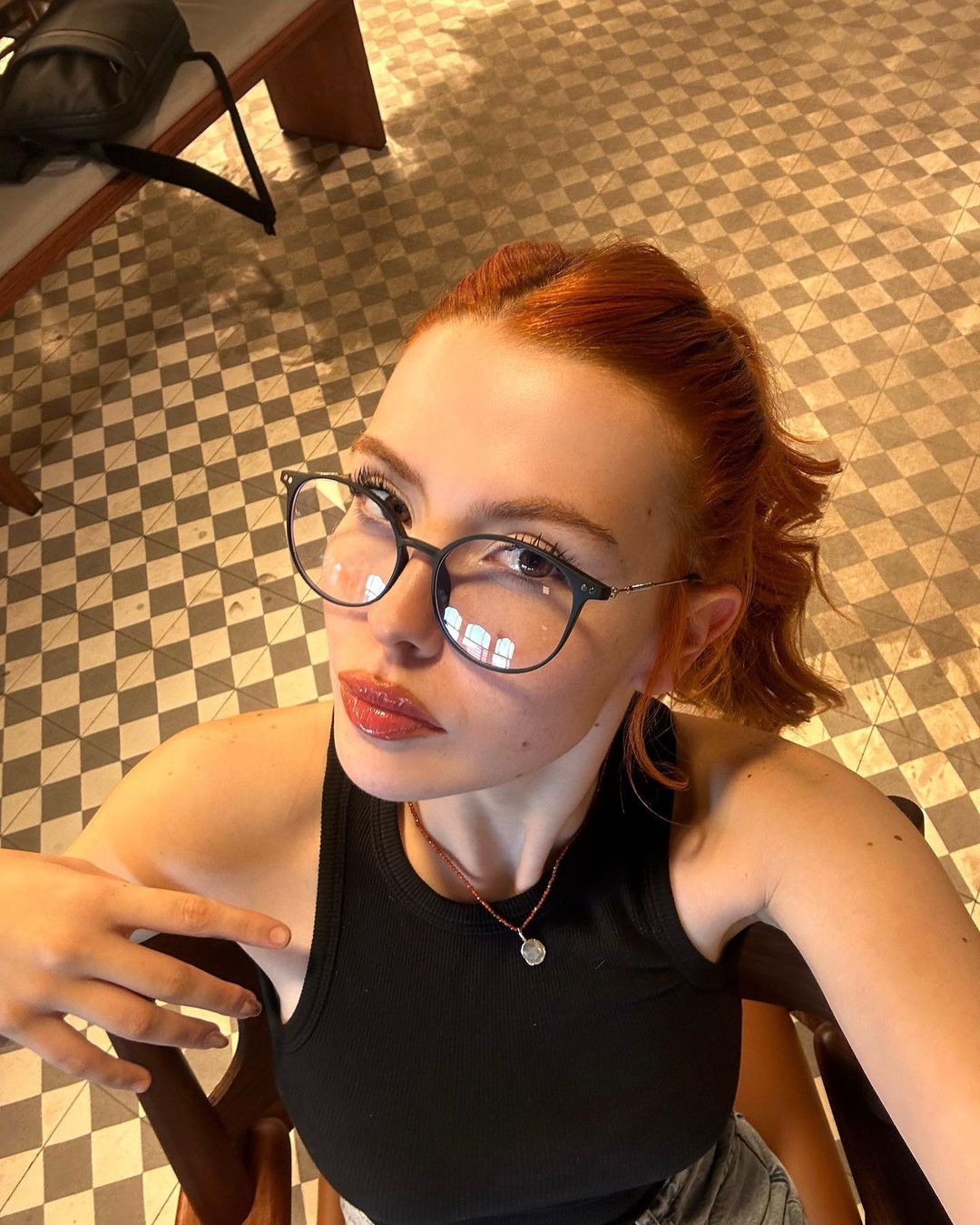 A person with red hair and glasses seated in a classic setting with geometric floor tiles.